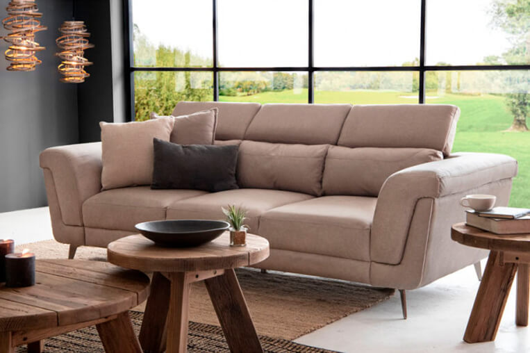 Three seater sandstone couch with rustic wooden sidetables in living room with large window overlooking a garden